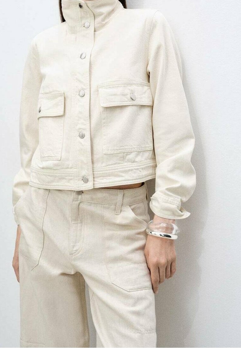Personne portant une veste en jean beige clair et un pantalon assorti, avec des bracelets en argent au poignet, debout devant un mur blanc uni.