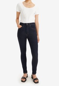 Vaqueros pitillo azul oscuro de talle alto, diseño de cinco bolsillos y costuras naranjas, combinados con una camiseta blanca de manga corta ajustada.