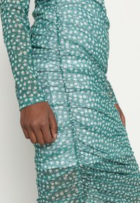 Robe florale verte en matériau transparent, dotée de manches longues, de fronces sur les côtés et d'une silhouette ajustée ornée de petites fleurs blanches et lavande.