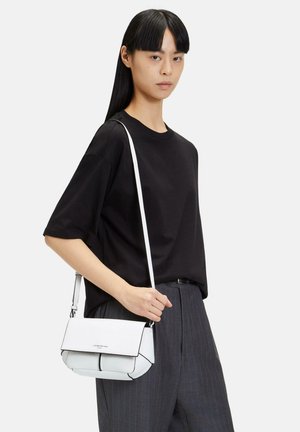 Sac en cuir blanc porté en travers, avec une surface texturée, des accents noirs et une sangle réglable, tenu par une personne portant un t-shirt noir.