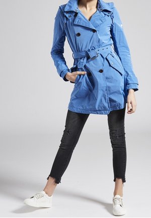 Trench-coat bleu léger avec un design croisé à double boutonnage, ceinture ajustable et détails de boutons. Associé à un jean noir déchiré et des baskets blanches.