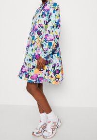 Blommig skjortklänning med blå bakgrund, prydd med livfulla lila, rosa och gula blommor. Långa ärmar och utsvängd nederkant. I sällskap med vita strumpor och sneakers med silverkant.