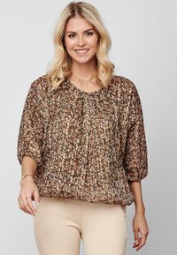 Smilende kvinde med blont hår iført en brun blomsterbuket og beige bukser, stående mod en lys grå baggrund.