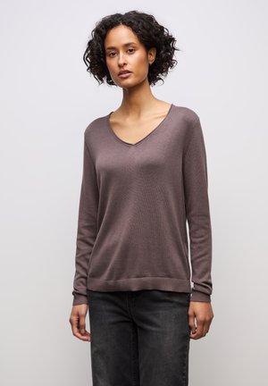 Ung kvinna med lockigt hår, klädd i en brun tröja med V-ringning och svarta jeans, stående mot en enkel ljusgrå bakgrund.