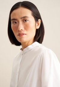 Camicia bianca con colletto e chiusura a bottoni, caratterizzata da leggerette maniche a sbuffo e un tessuto liscio e leggermente trasparente. Design semplice ed elegante.
