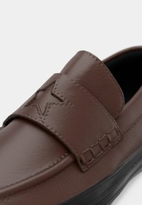 Pantofi slip-on din piele maro, cu un design cusut, accente în formă de stea și o talpă din cauciuc negru. Textură netedă cu un aspect minimalist.
