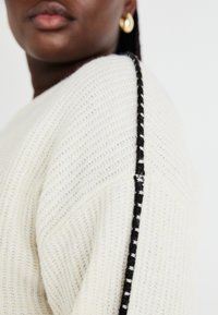 Κρεμ μανίκι πλεκτό πουλόβερ με υφασμάτινη υφή, πλαϊνές λεπτομέρειες σε μαύρο και άσπρο ρίγα και χαλαρή εφαρμογή. Διακοσμημένο με ριμπά πλάγια.