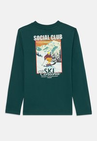 Zöld, hosszú ujjú póló, síelőt, hegyeket és fákat ábrázoló grafikával, rajta a "SOCIAL CLUB" és a "SKI Cortina" felirattal.