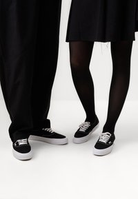 Des baskets noires en toile avec des lacets blancs et des semelles en caoutchouc blanches, associées à des vêtements sombres et des collants noirs, sur fond blanc.