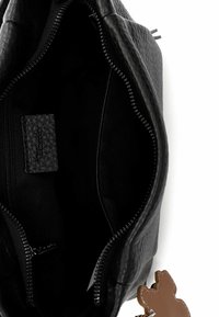 Musta teksturoitu nahkainen käsilaukun sisäosa, jossa on vetoketjusuljenta, sileä musta vuori ja siihen kiinnitetty ruskean karhunmuotoinen charmikoriste.