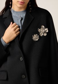 Schwarzer Mantel mit zwei metallischen Blumenbroschen, eine größere mit einem zentralen Kristall, die andere kleiner. Darunter ein grauer, strukturierter Pullover.