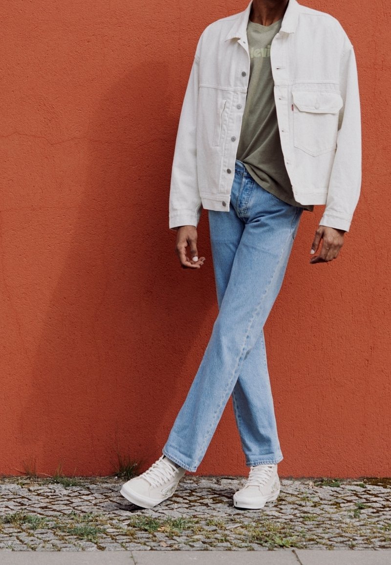 Osoba v bílém kabátu, zeleném tričku, modrých džínách a bílých teniskách, stojící s překříženýma nohama proti pozadí oranžové zdi.