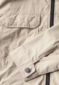 Beige lehká bunda se zipem vpředu, náprsní kapsou a rukávem na knoflík. Hladká textura a minimální detaily šití.