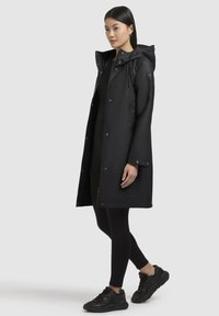 Zwarte waterdichte jas met capuchon, voorzien van een rits aan de voorkant en drukknoopsluitingen, gedragen over zwarte leggings en sneakers. Glad materiaal, lange snit.
