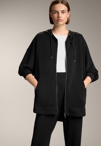 Felpa nera con zip e vestibilità morbida, dotata di cappuccio, due tasche frontali e polsini a costine. Abbinata a pantaloni neri coordinati.