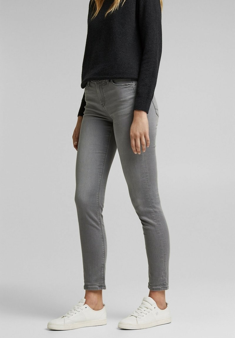 Femme portant un jean skinny gris, un haut à manches longues noir et des baskets blanches, debout devant un fond gris uni.