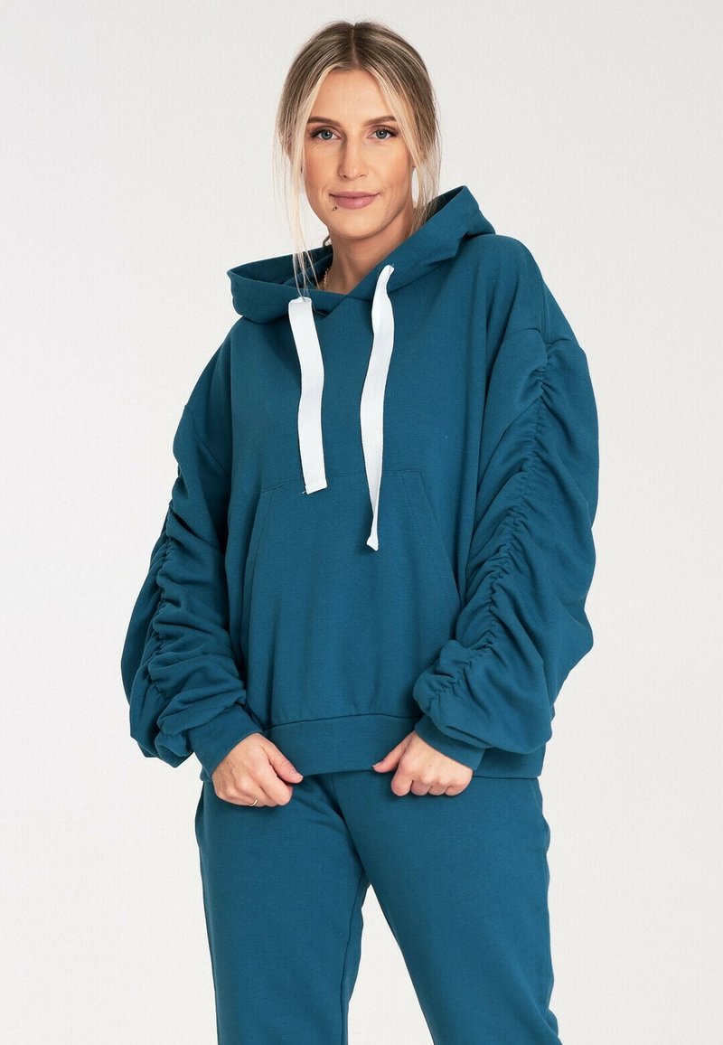 Turkusowa bluza z kapturem o luźnym kroju, z białymi sznureczkami i marszczonymi rękawami. Posiada przednią kieszeń oraz ściągacze na mankietach.