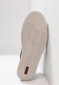Scarpa nera con suola in gomma color crema caratterizzata da un motivo a zigzag e un accento rosso del logo sul lato. Vista dal basso su uno sfondo bianco.