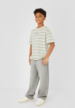 Junge mit kurzen lockigen Haaren steht lächelnd da, trägt ein beiges gestreiftes T-Shirt, lockere graue Jeans und weiße Turnschuhe vor einem schlichten Hintergrund.