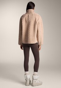 Mehka bež jakna z vzorcem kabelskega pletenja, visokim ovratnikom in kratkim krojem, kombinirana z rjavimi pajkicami in svetlimi zimskimi škornji.