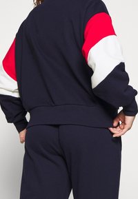 Felpa navy con righe di accento rosse e bianche sulle maniche. Texture morbida, orlo a coste, vestibilità rilassata, con una tasca visibile.