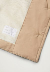 Pehmeä, kermaisen värinen tekoturkiskangas yhdellä puolella, beige satiinimainen ulkopinta, jossa on ommeltu paneeli. Mukana valkoinen kangastunnus nimelle ja puhelintiedoille.