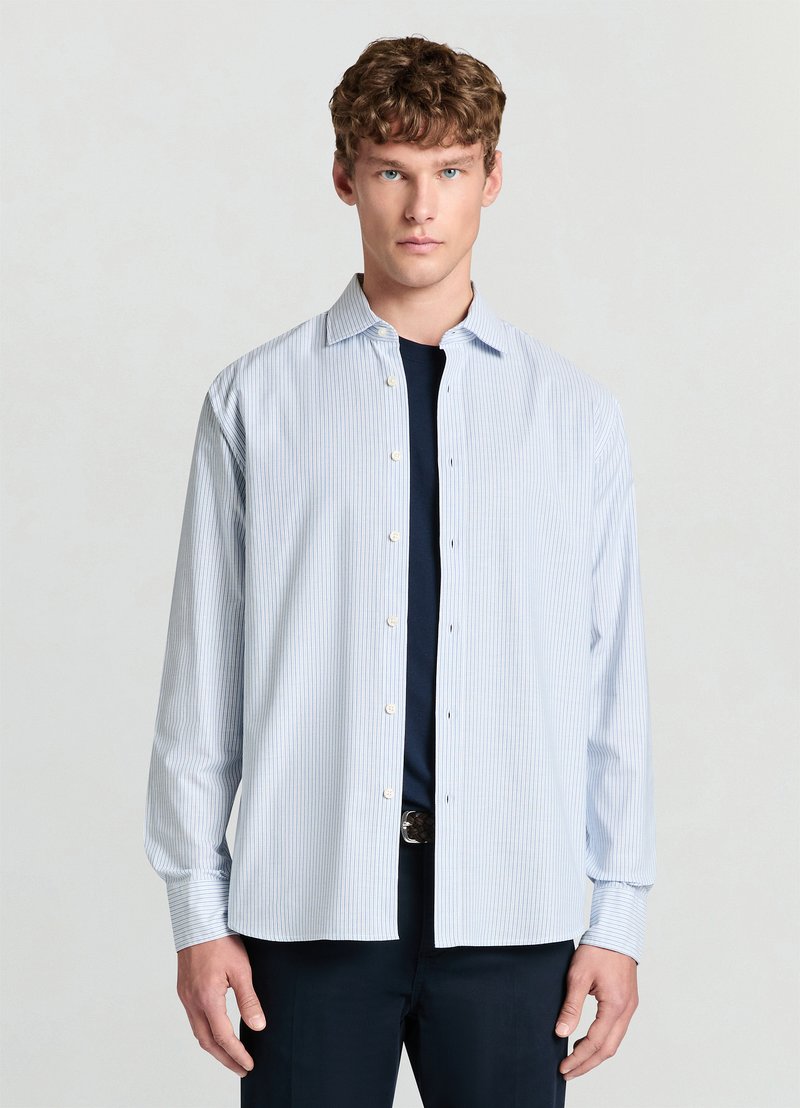 Camicia a righe azzurro chiaro con collo button-down, maniche lunghe e dettagli discreti sui polsini, abbinata a una maglia blu scuro sotto.
