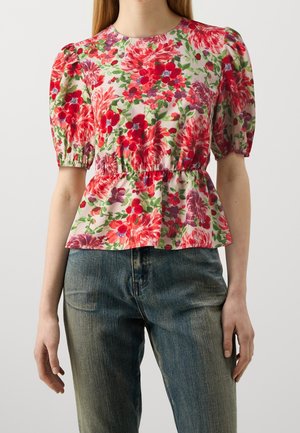 Kvinde iført en bluse med blomsterprint og puffede ærmer i røde, pink og lilla nuancer, kombineret med blå denimjeans mod en ensfarvet baggrund.