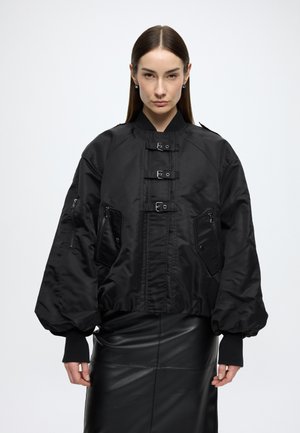 Fekete bomber dzseki szatén anyagból, elöl cipzárral, nagy zsebekkel és bordázott mandzsettákkal; széles, rövidített sziluettel.