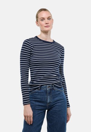 Femme aux cheveux blonds portant un shirt à manches longues rayé bleu marine et blanc et un jean bleu taille haute, debout devant un fond uni.