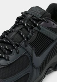 Gros plan sur une chaussure Nike Zoomero 5 noire présentant un mesh texturé, des lacets noirs réfléchissants et un grand swoosh emblématique sur le panneau latéral.