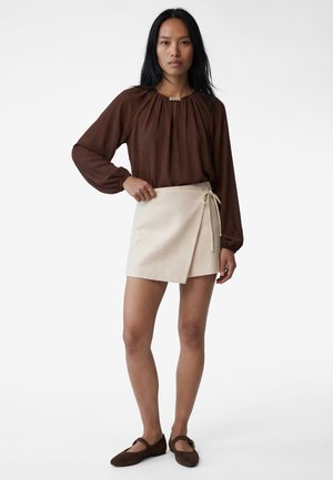 Donna in piedi con la mano sulla vita, indossa una blusa marrone a maniche lunghe, gonna avvolgente beige e scarpe basse marrone scuro su sfondo bianco.