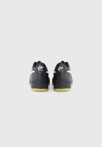 Crne Nike Air tenisice s glatkom kožnom vanjštinom, bijelim Swoosh detaljima i žutom gumiranom potplatnom, prikazane sa stražnje strane.