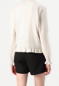 Suéter de punto color beige con un corte relajado, que presenta un dobladillo y puños acanalados. Superficie texturizada con un patrón sutil. Combinado con pantalones cortos negros.