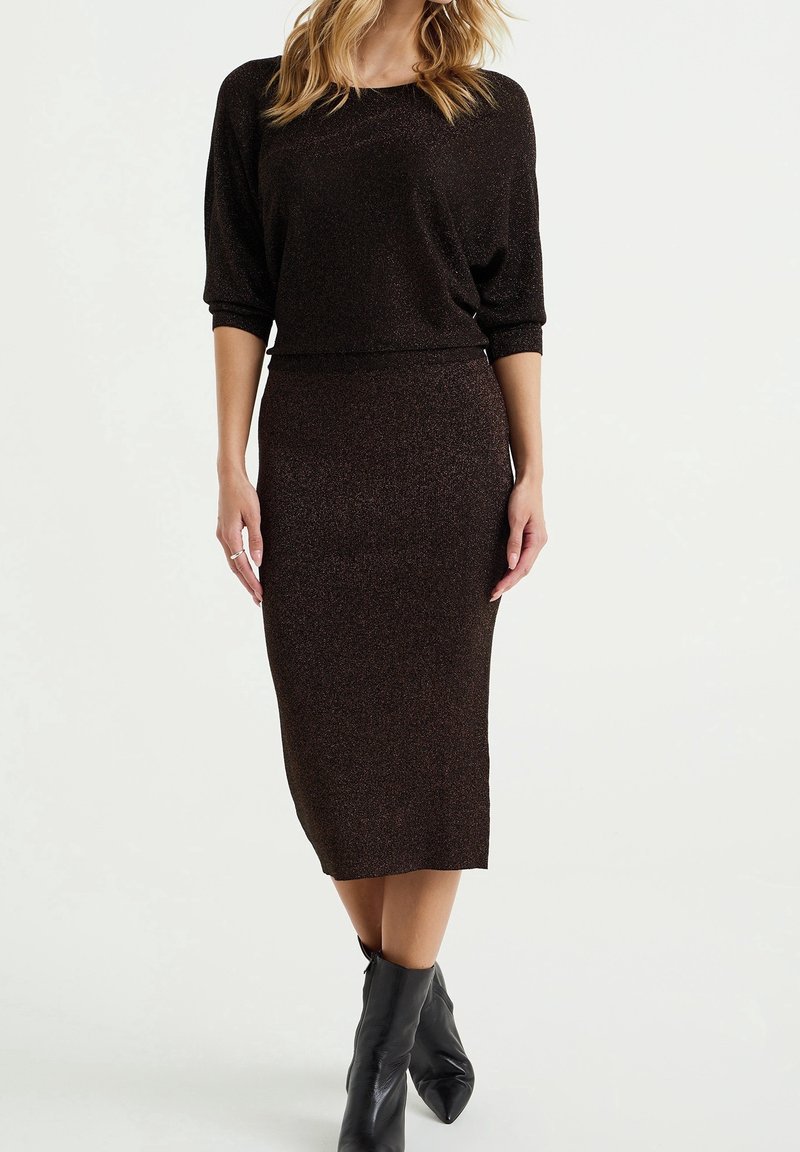 Femme portant une tenue deux pièces marron foncé scintillante avec une jupe midi ajustée et un top à manches trois-quarts, associée à des bottines noires.