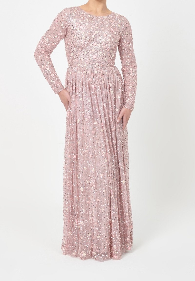 Robe rose à manches longues et longueur jusqu'au sol, ornée de paillettes florales, portée par une personne debout devant un fond blanc.