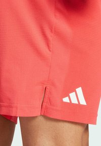 Shorts de sport corail en tissu texturé, dotés d'une fente sur le côté et du logo Adidas blanc avec trois bandes diagonales.