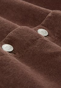 Bruine, gestructureerde stof met zichtbare zilveren knopen. Het materiaal lijkt zacht en pluizig, met zichtbare stiksels en een gevouwen ontwerp.