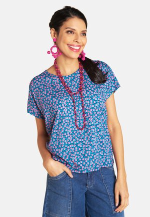 Frau lächelt, trägt ein blau gemustertes Oberteil, Jeanshose, große rosa Perlenohrringe und eine passende lange rosa Perlenkette, Hand in der Tasche.