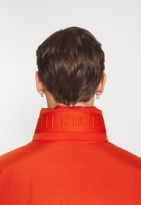 Bakåtsikt av en person med kort brunt hår som bär en röd jacka med "HILFIGER" broderat på kragen, silverfärgade hoop-örhängen synliga.