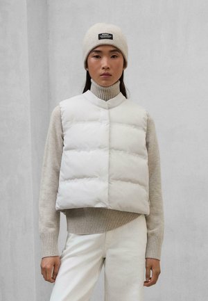 Chaleco blanco esponjoso con un cuello alto, con cierre de botones y patrón acolchado. Lleva puesto un suéter de cuello alto beige claro y pantalones claros.