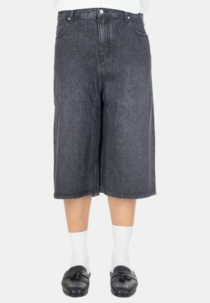 Persona che indossa pantaloncini neri in denim larghi fino al ginocchio, calzini bianchi e mocassini in pelle neri con nappine su sfondo bianco.