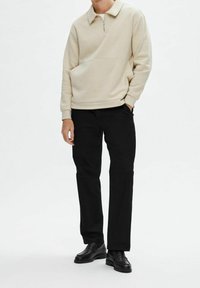 Man i beige kvartdraget tröja, svarta byxor och svarta läder loafers, stående med ena handen i fickan mot en enfärgad bakgrund.