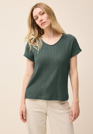 Donkergroen T-shirt met korte mouwen van een gestructureerde stof en een ronde halslijn, gecombineerd met lichtgekleurde broeken. Eenvoudige, losse pasvorm.