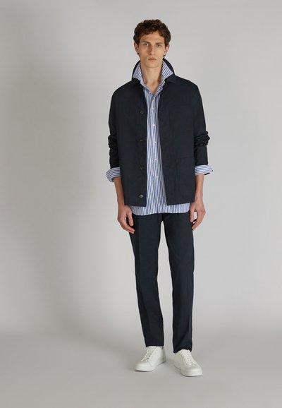 Chaqueta azul marino oscura con un alto cuello, detalle de botones y un bolsillo de parche sobre una camisa de rayas azul y blanca, combinada con pantalones oscuros y zapatillas blancas.