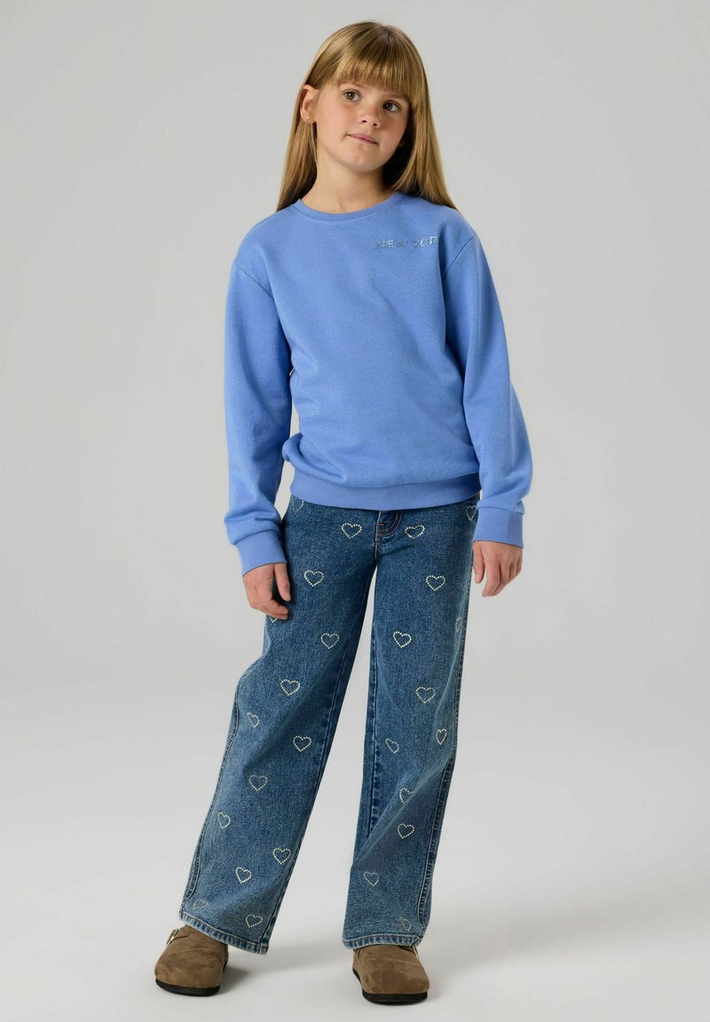 Felpa azzurro chiaro con scollo rotondo e scritta "NEW YORK"; abbinata a jeans a gamba larga con motivi a forma di cuore e calzature color cuoio.