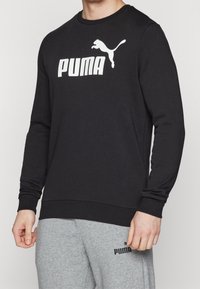 Hombre con sudadera de manga larga negra de Puma y pantalones de chándal grises claros de Puma, de pie con los brazos relajados contra un fondo plano.