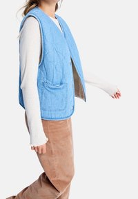 Lichtblauwe gewatteerde bodywarmer met een textuurpatroon, voorzakken en drukknoopsluitingen, gedragen over een wit shirt met lange mouwen en bruine ribfluwelen broek.