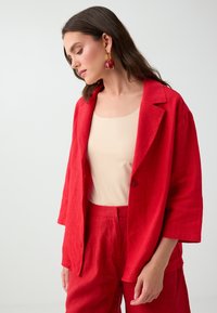 Femme aux longs cheveux bruns portant un blazer rouge et un pantalon assorti sur un haut beige, regardant vers le bas sur un fond uni.