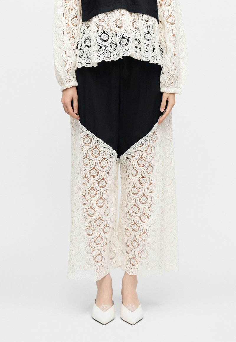 Femme portant un pantalon noir avec des panneaux larges en dentelle blanche et un haut assorti en dentelle, assortis à des chaussures blanches à bout pointu.